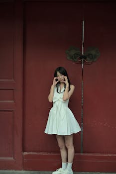 Stylish young woman in white dress posing playfully against a large red door.