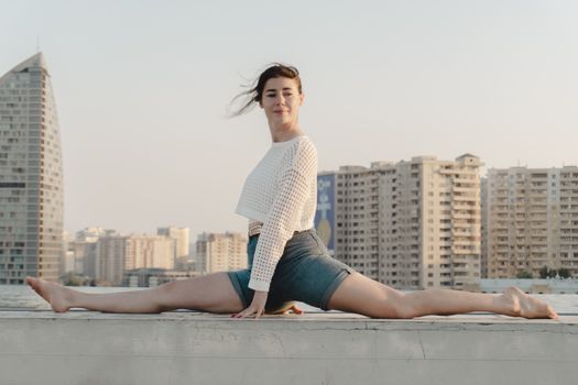 A young woman performs a splits exercise on a rooftop, with a city skyline backdrop.