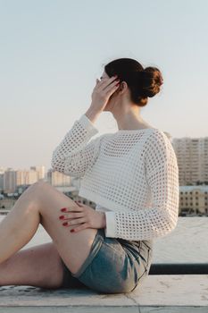 Stylish woman in white top and denim shorts sitting on rooftop with cityscape backdrop.