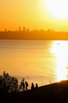 Golden sunset illuminating city skyline across calm water with silhouettes of people by the shore.