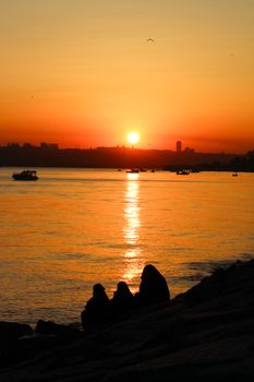 Silhouetted figures sit by the calm water under a mesmerizing sunset sky.
