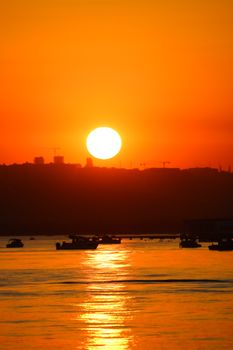 A stunning golden sunset over a serene water body with silhouetted boats and skyline.