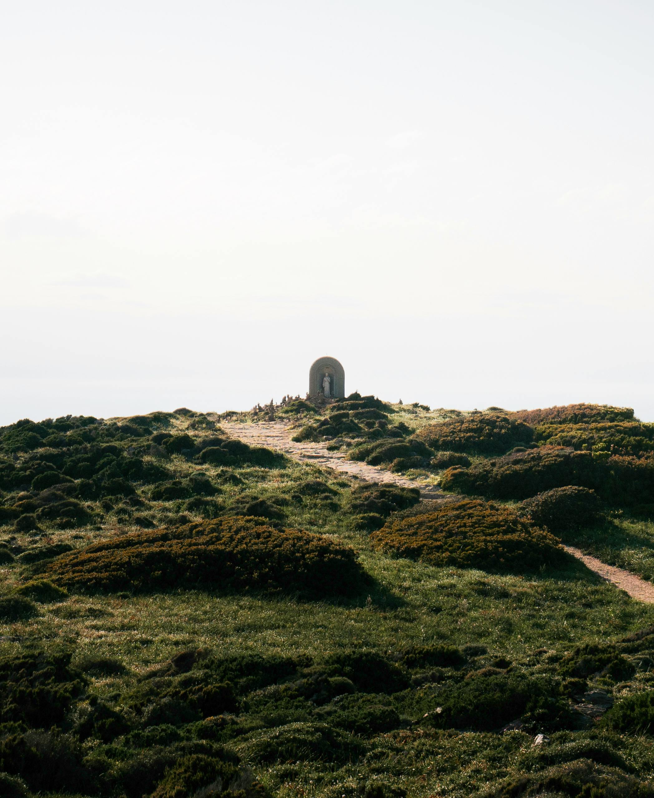 Explore a tranquil pathway leading to a memorial on a Corsican hillside with lush greenery.