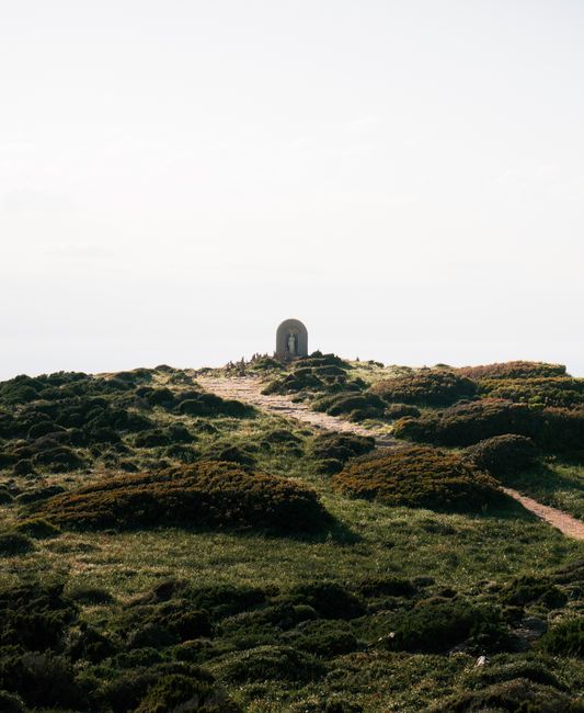 Explore a tranquil pathway leading to a memorial on a Corsican hillside with lush greenery.