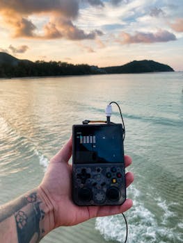 A person holding a retro gaming console at sunset on a scenic beach.