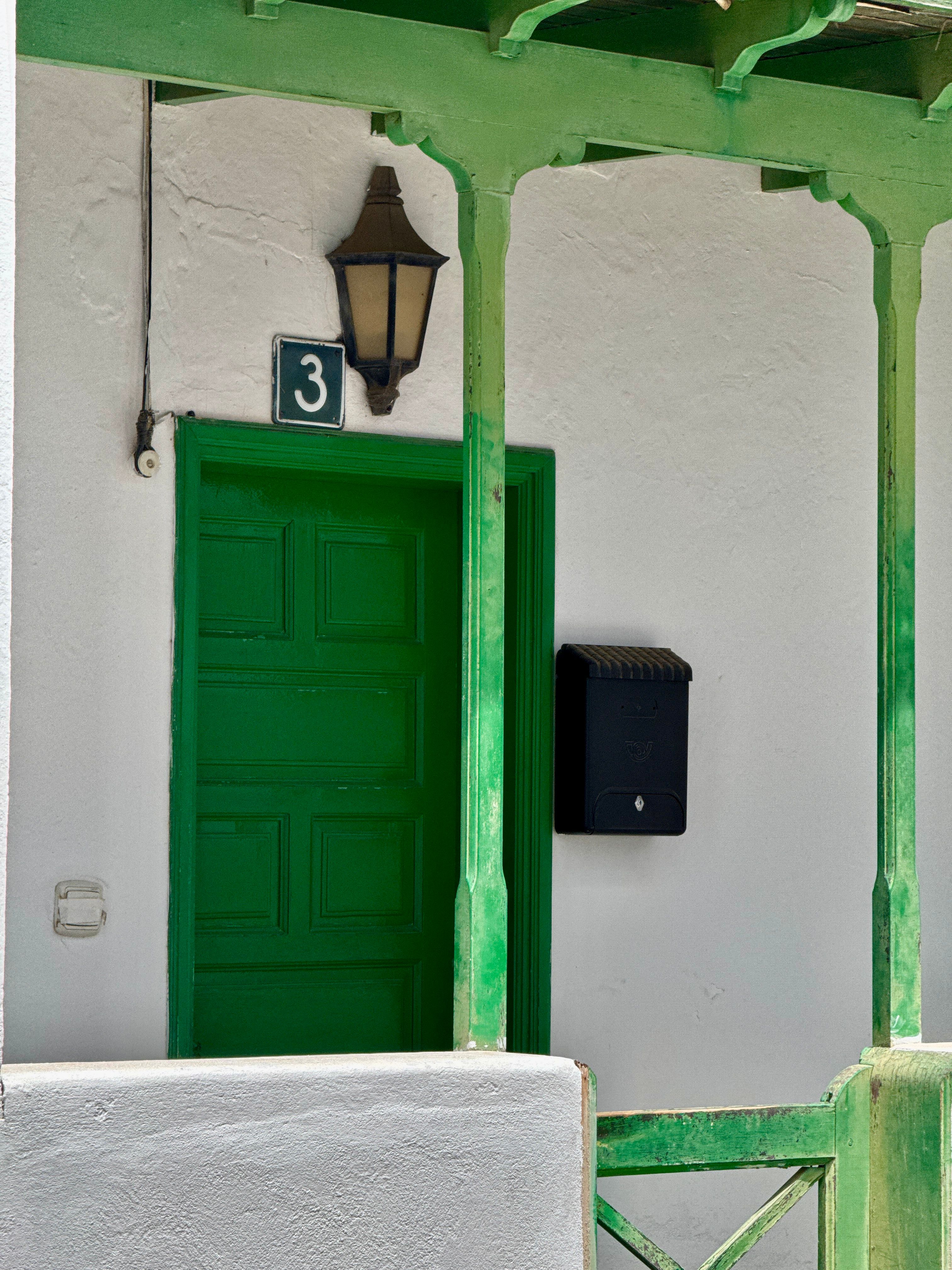 A vibrant green door and mailbox on a white wall in a traditional Lanzarote street, under a vintage lantern.