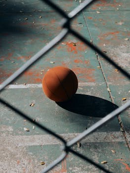 A basketball on an outdoor court viewed through a chain-link fence, highlighting urban sports.