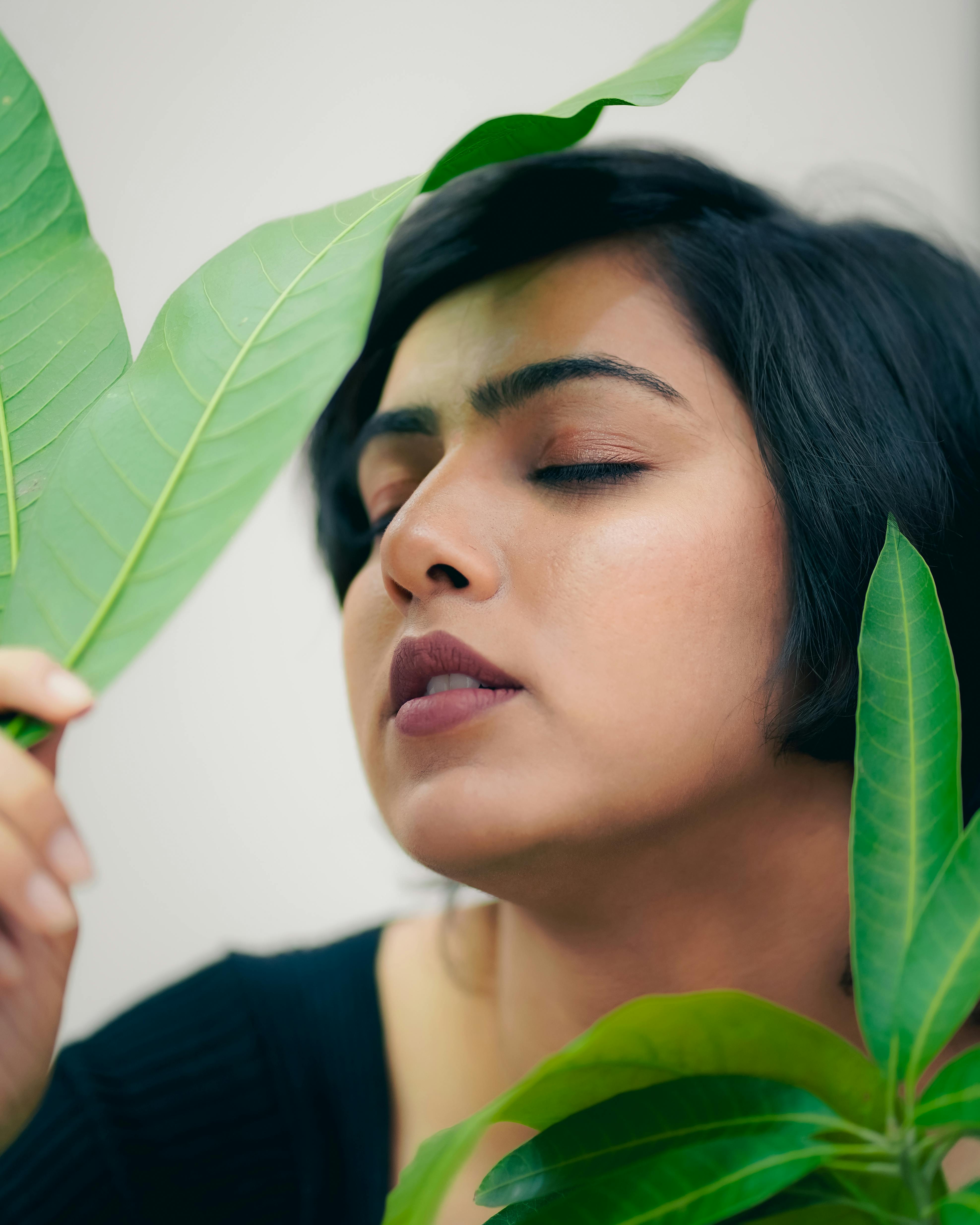 Close-up portrait of a woman surrounded by green leaves, eyes closed.