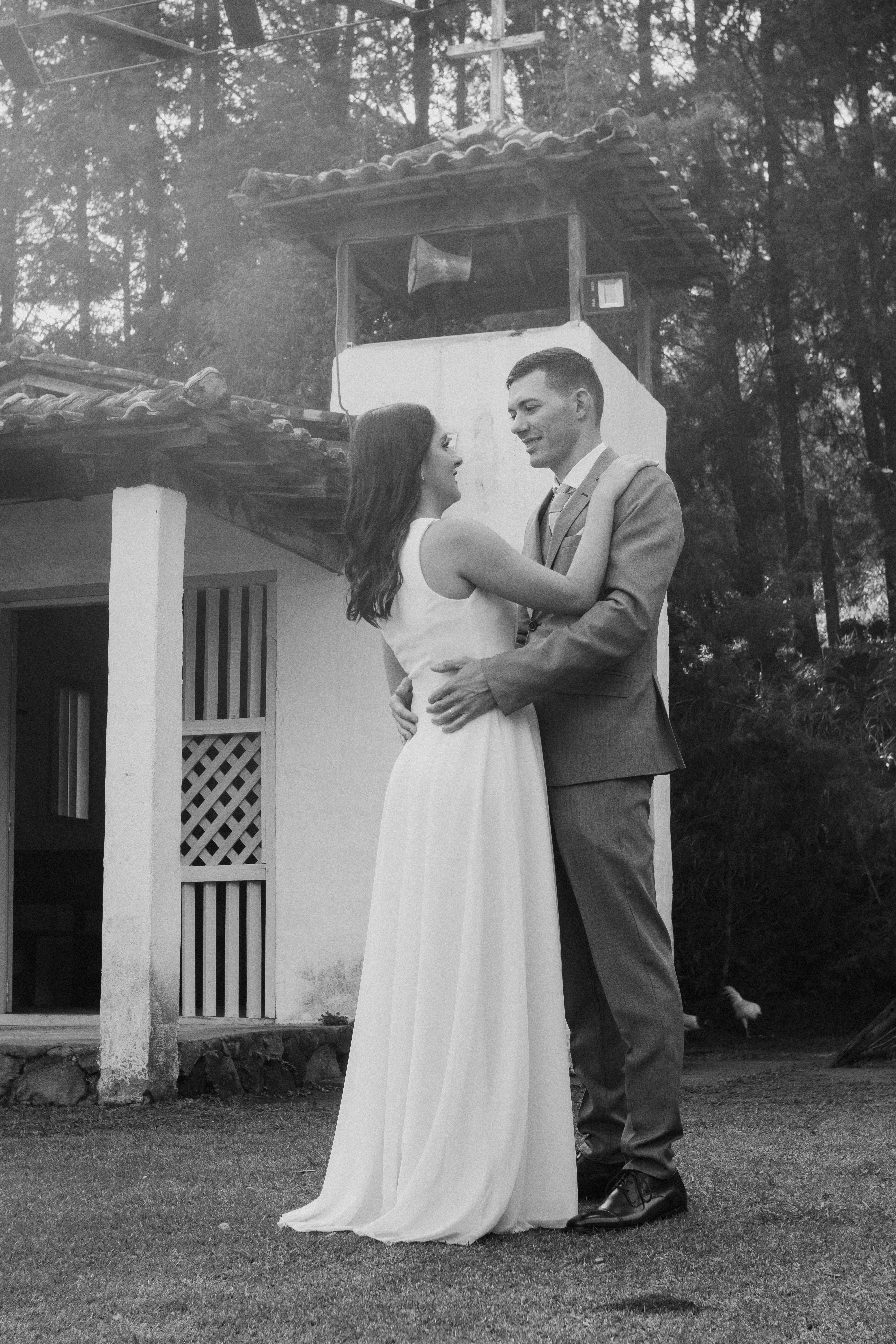 Newlywed couple embraces in front of rustic chapel in forest setting.