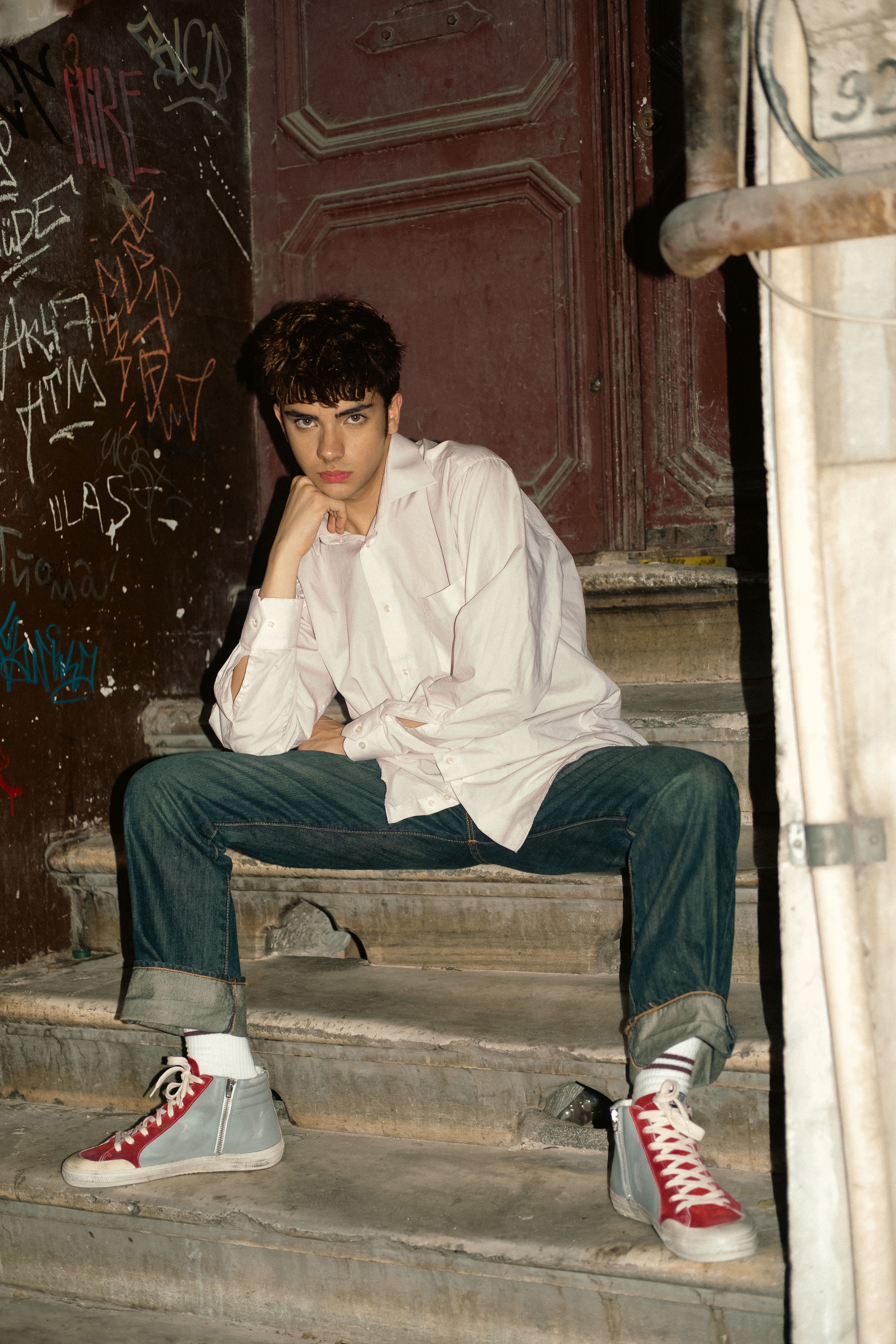 Stylish young man posing on steps in İstanbul with an urban backdrop, showcasing modern fashion trends.