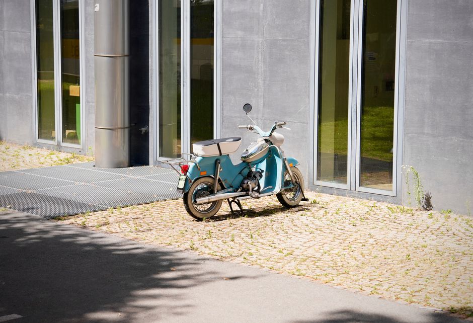 Retro blue scooter parked outside a modern glass building under sunlight.