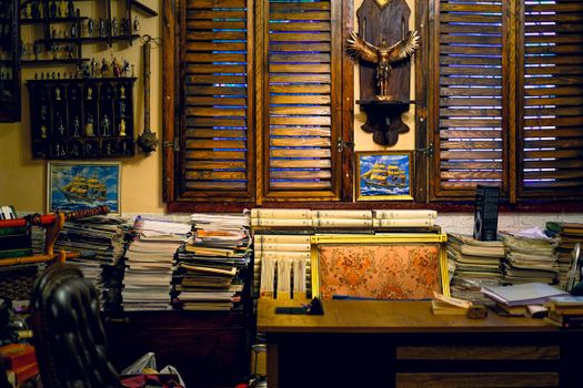Warm, cluttered study room featuring vintage decor, wooden blinds, and stacks of papers and books.