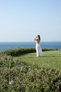 A woman in a white skirt admires the ocean from a grassy cliff on a clear, sunny day.