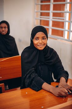 Portrait of a young woman in a hijab sitting in a classroom, smiling warmly.