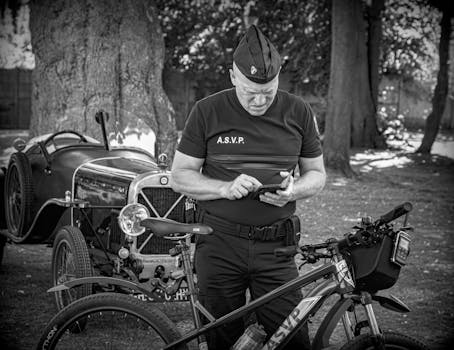 Man in uniform checks device near vintage car and bicycle in park setting, black and white photo.