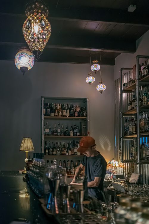 Free Bartender working in a dimly lit bar with decorative lamps and shelves of bottles. Stock Photo