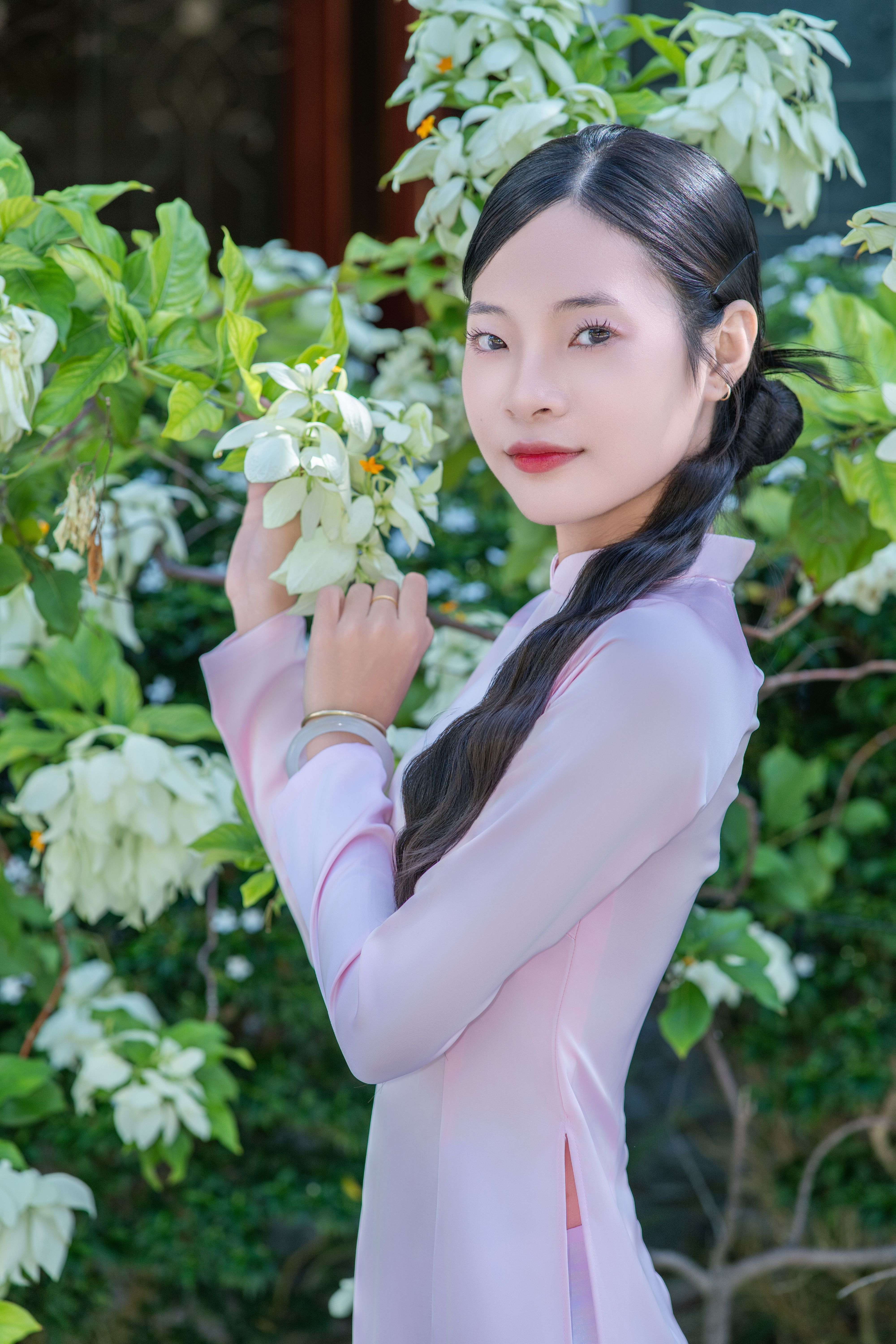 Woman in traditional Ao Dai dress poses among blooming white flowers outdoors.