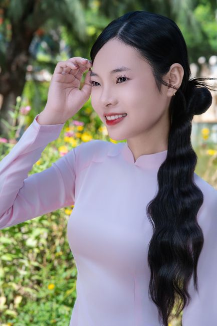 Asian woman in pink dress smiling outdoors, surrounded by greenery.