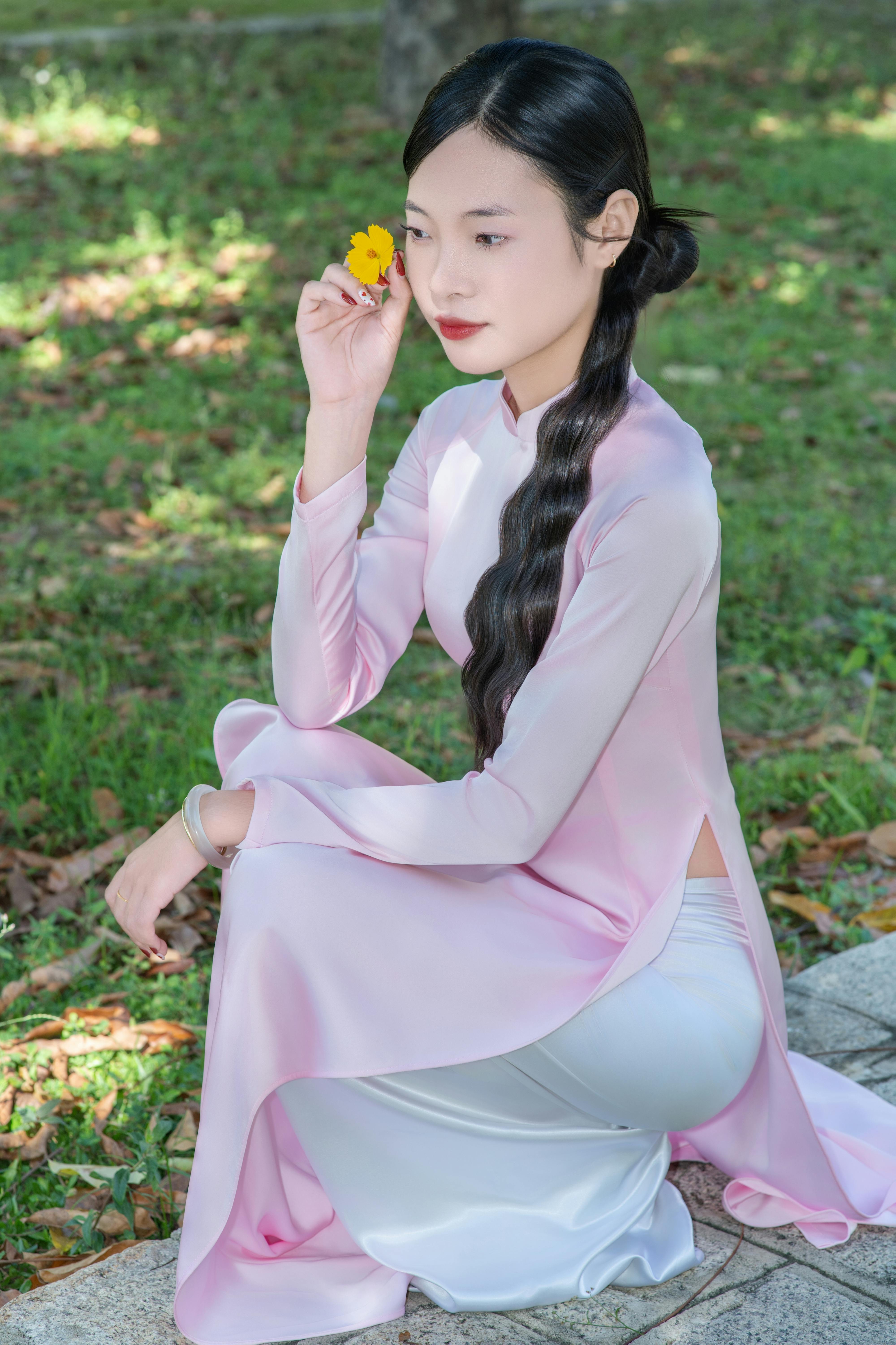 A woman in pink ao dai holding a yellow flower, seated on a sunny day outdoors.