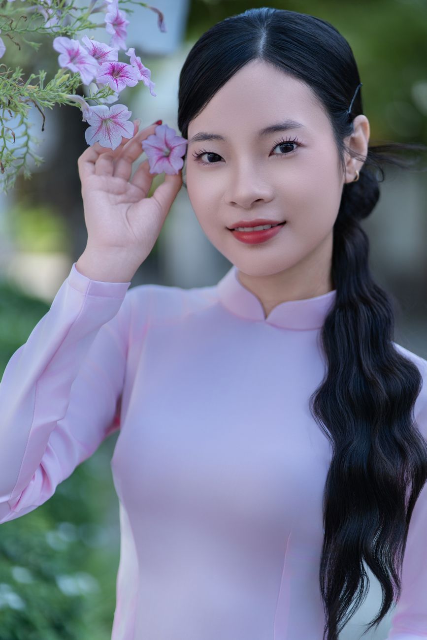 Young woman in traditional pink Ao Dai holding pink flowers outdoors, smiling.