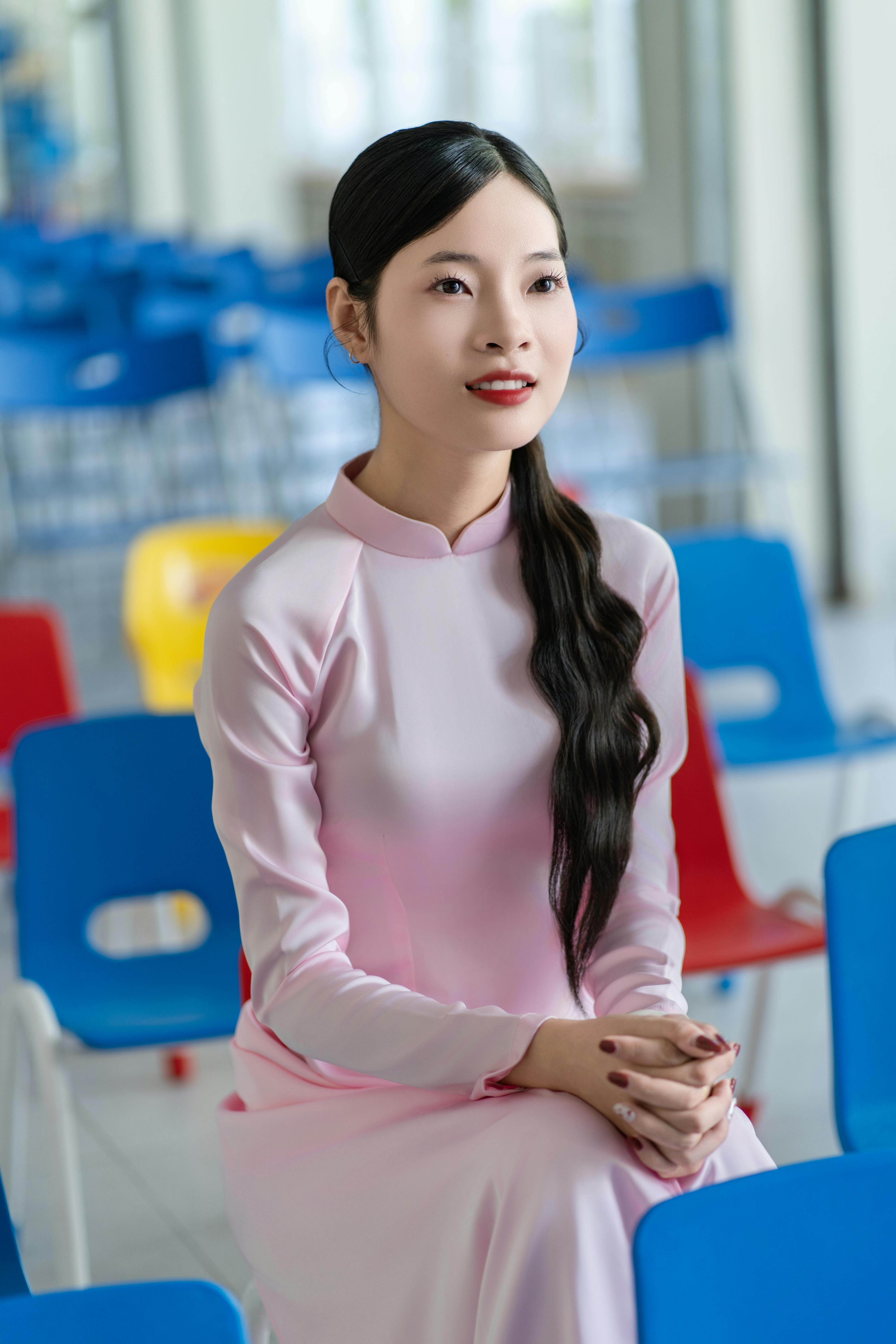 A young woman wearing a pink Ao Dai sits in a colorful, bright room, exuding elegance.