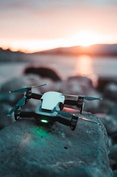 A drone sits on rocky terrain with a vibrant sunset reflecting over water.