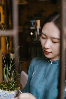 A young woman in a traditional dress lovingly gazes at a potted plant indoors.