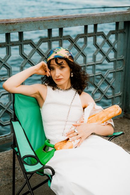 Stylish woman sitting by riverside holding a baguette. Urban tranquility captured.