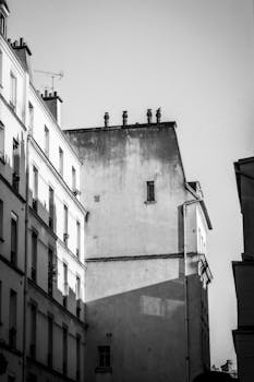 Black and white photo of Parisian buildings with dramatic shadows, evoking classic urban charm.