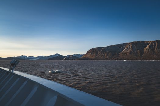 A serene view of mountains and icebergs from a ship in the Arctic Ocean at sunset.