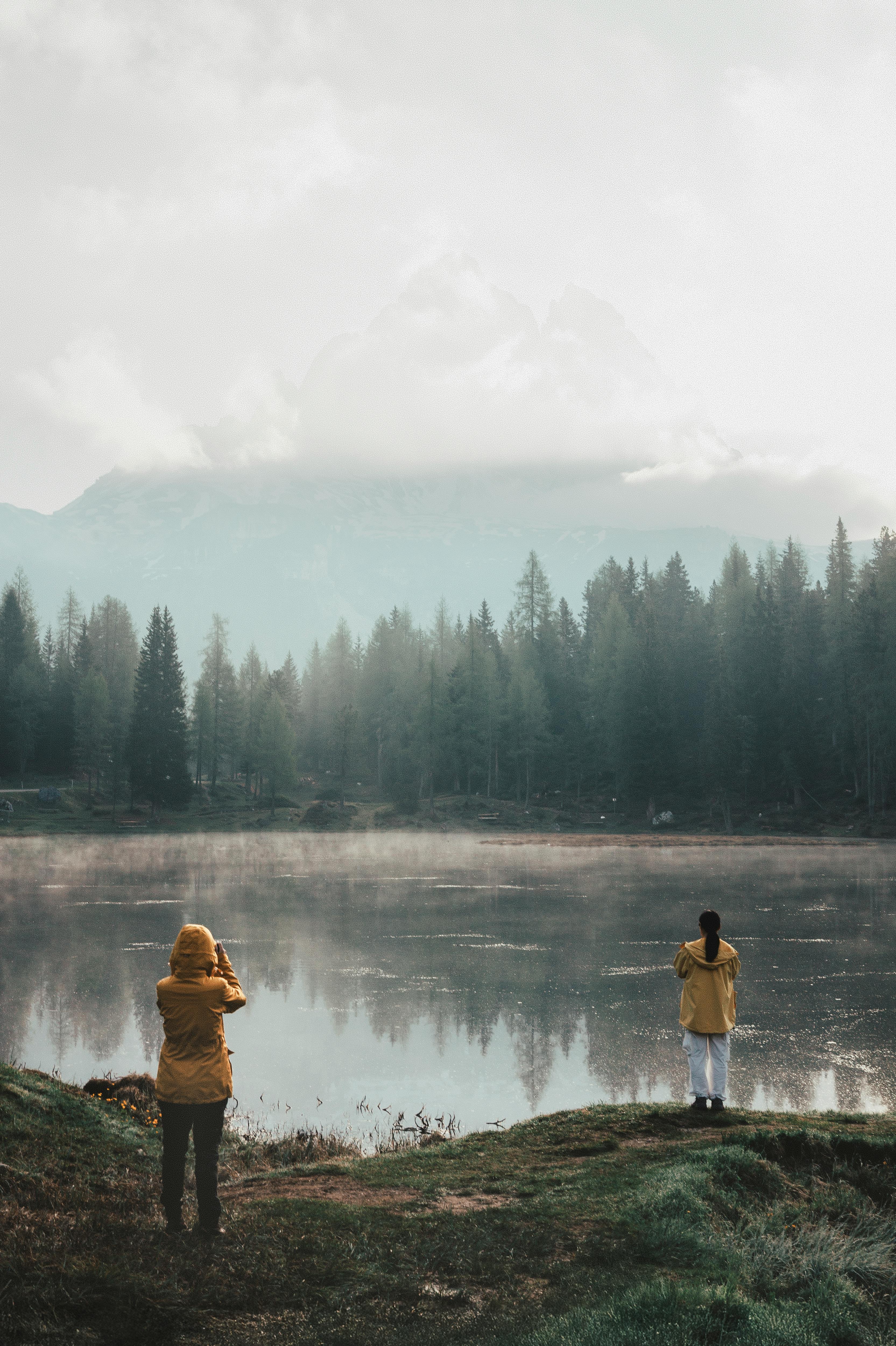 Two hikers in yellow jackets admire a foggy mountain lake view, surrounded by pine trees.
