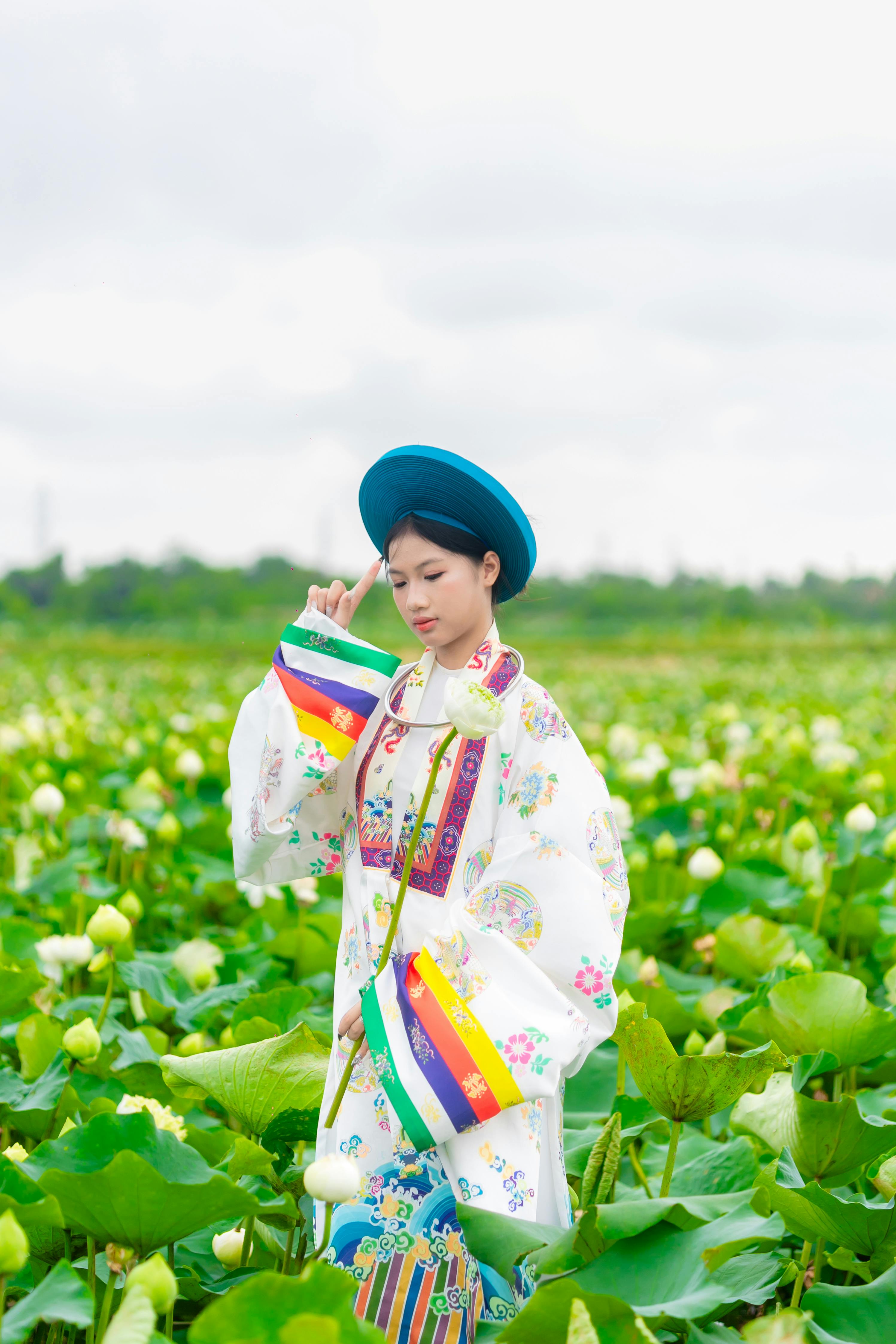 A young woman in vibrant traditional attire stands amidst a lush lotus field under a cloudy sky.