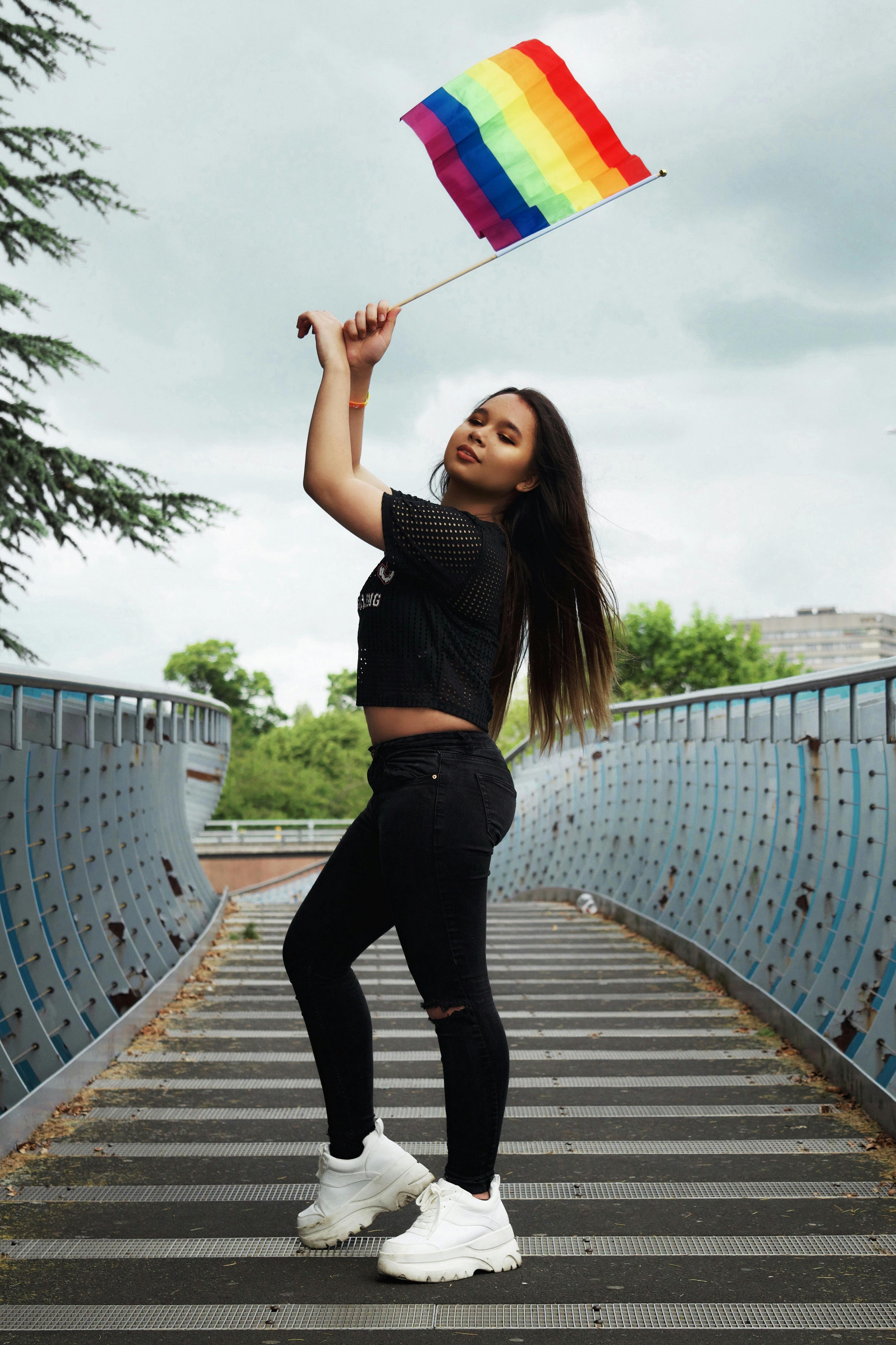 一位穿着黑色服装的自信年轻女子站在桥上，挥舞着彩虹旗，在户外庆祝骄傲