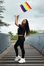 一位穿着黑色服装的自信年轻女子站在桥上，挥舞着彩虹旗，在户外庆祝骄傲