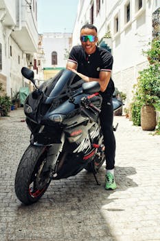 Man wearing sunglasses poses with a sleek motorcycle on a sunny day in a narrow alley.