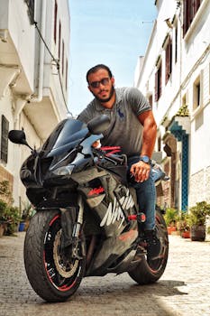 A man in casual attire leans on a sports motorcycle in a sunlit Mediterranean alleyway.
