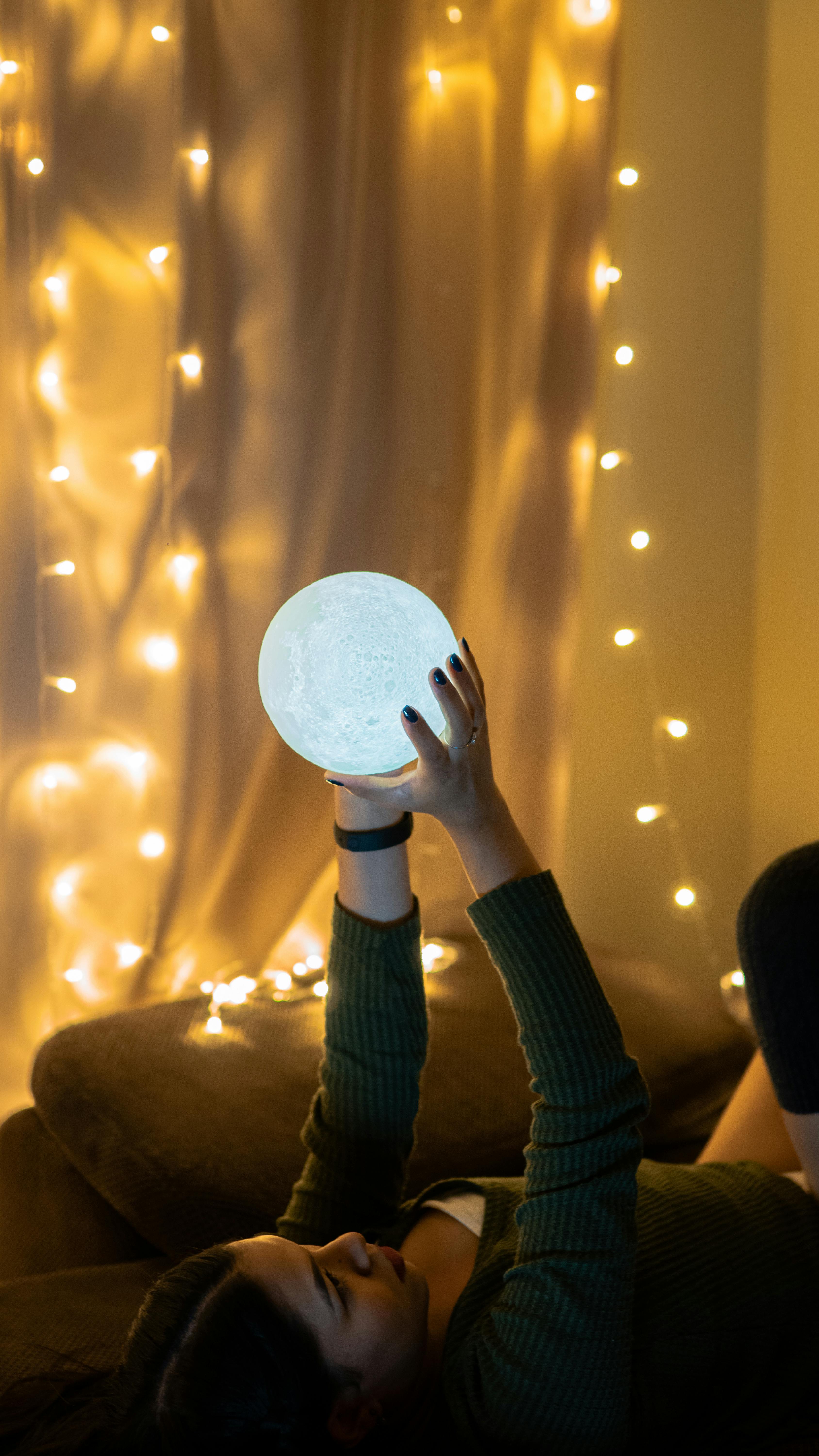 A woman relaxes indoors, enjoying the soft glow of a moon lamp and string lights.