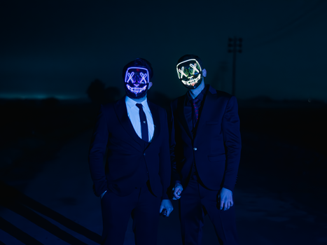 Two men wearing neon LED masks in dark suits, standing outdoors at night against a moody backdrop.