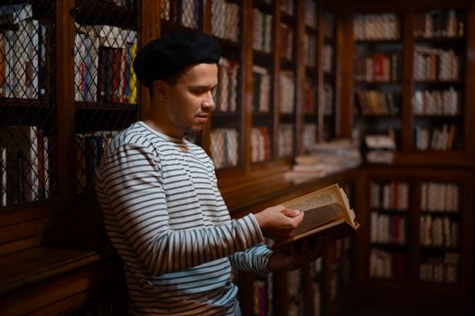 A man in a striped shirt reads a book in a warmly lit library filled with bookshelves.