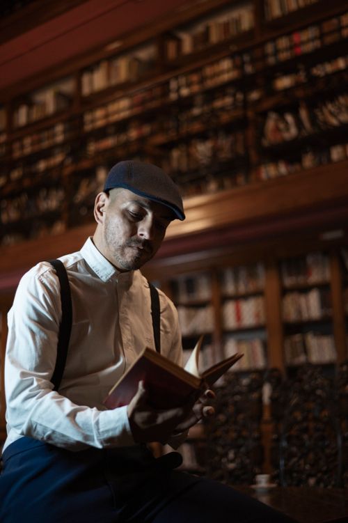 Free Adult man reading a book in a cozy library, wearing vintage clothing. Dimly lit, warm ambiance. Stock Photo