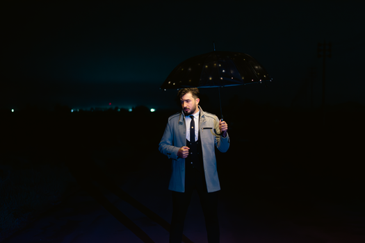 A stylish man in a suit and coat holds an umbrella under night lights, exuding style and charm.