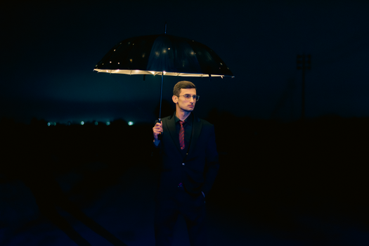 A man in a suit with glasses holding a glowing umbrella against a dark night background.