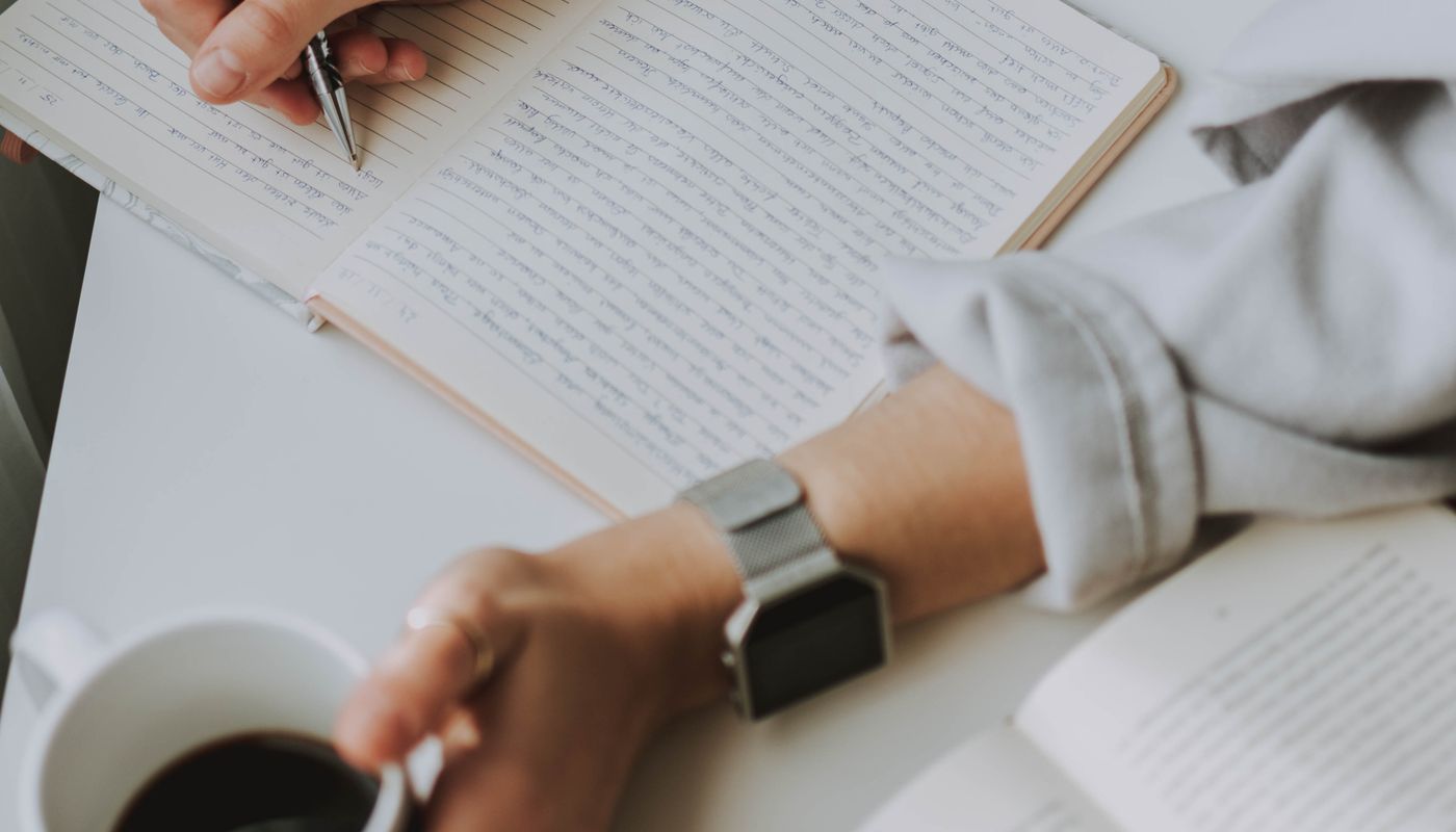 A person writing in a notebook at a desk while holding a coffee mug