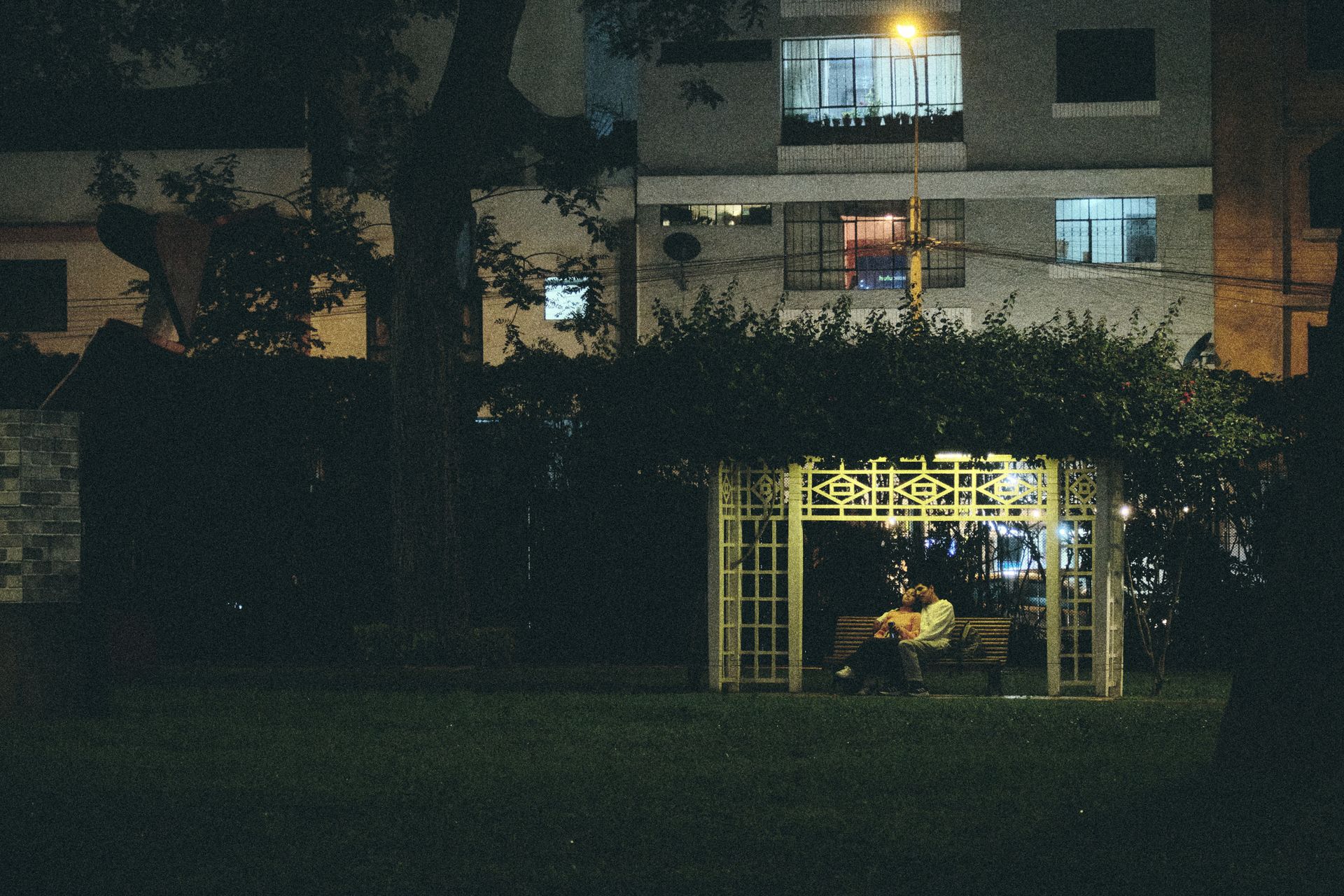 Couple sitting on a bench under a lit pergola in an urban garden at night.