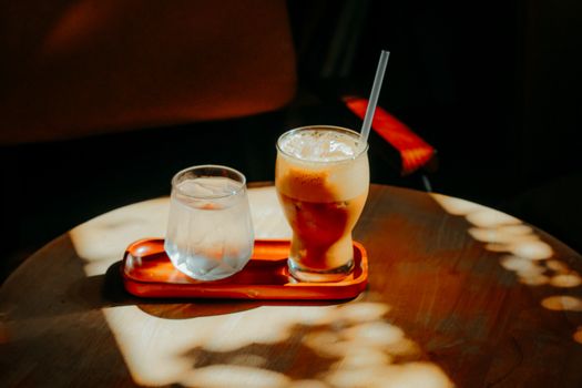 A glass of iced coffee and a glass of water on a tray, sunlight filtering through leaves.