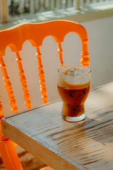 Glass of iced coffee with foam on a wooden table, sunlight through a bright chair.