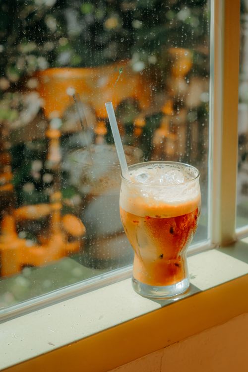 Free A glass of iced tea with a straw sits on a sunny window sill, casting warm reflections. Stock Photo