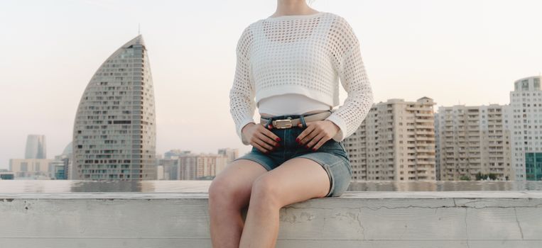 A woman in casual attire poses on a rooftop with a cityscape background during sunset.