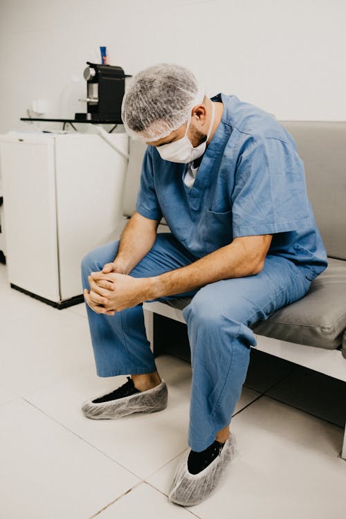 Man Wearing Blue Scrub Suit and Mask Sitting on Bench