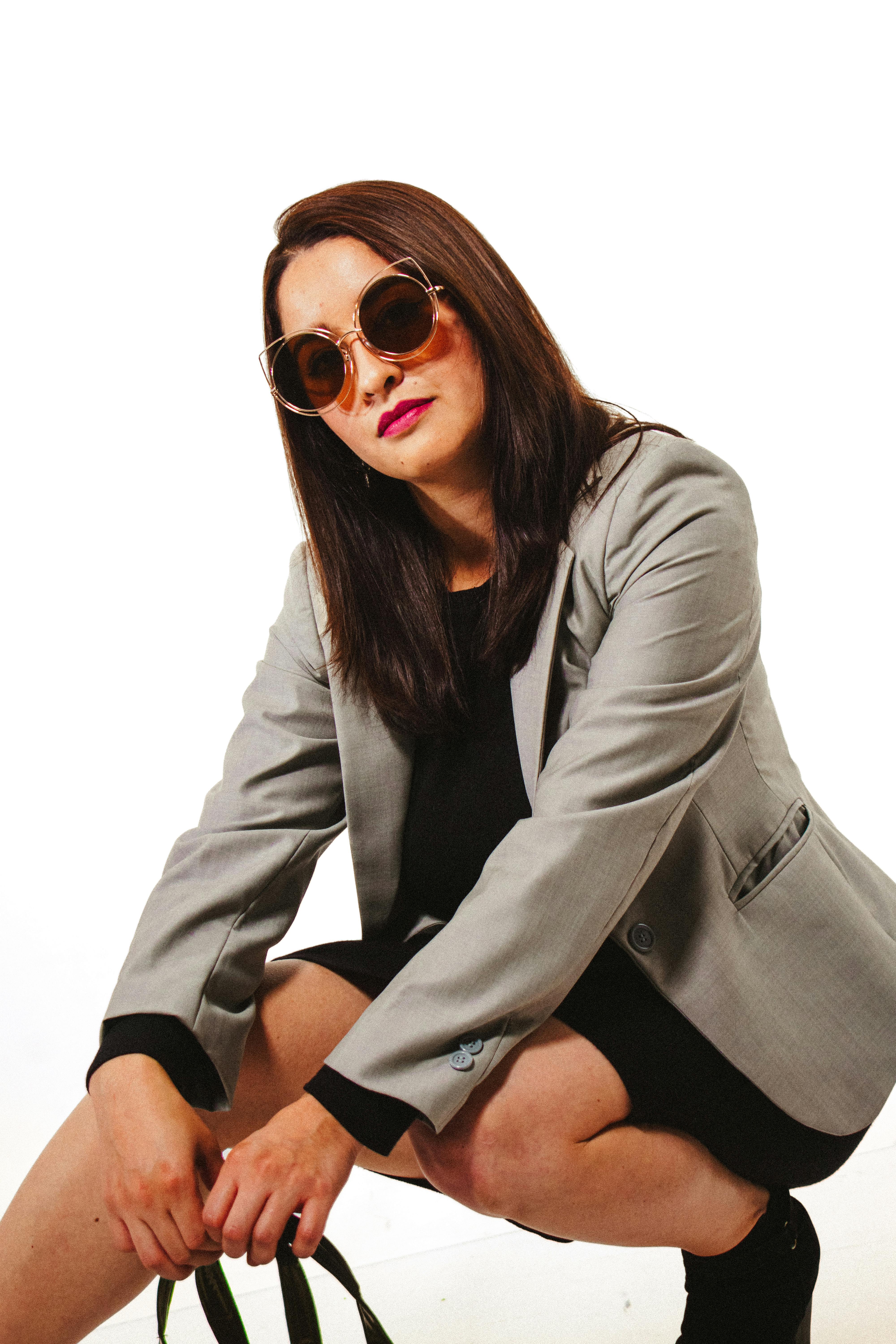 Gratis Una donna elegante che indossa un abito e occhiali da sole posa con sicurezza su uno sfondo bianco. Foto a disposizione